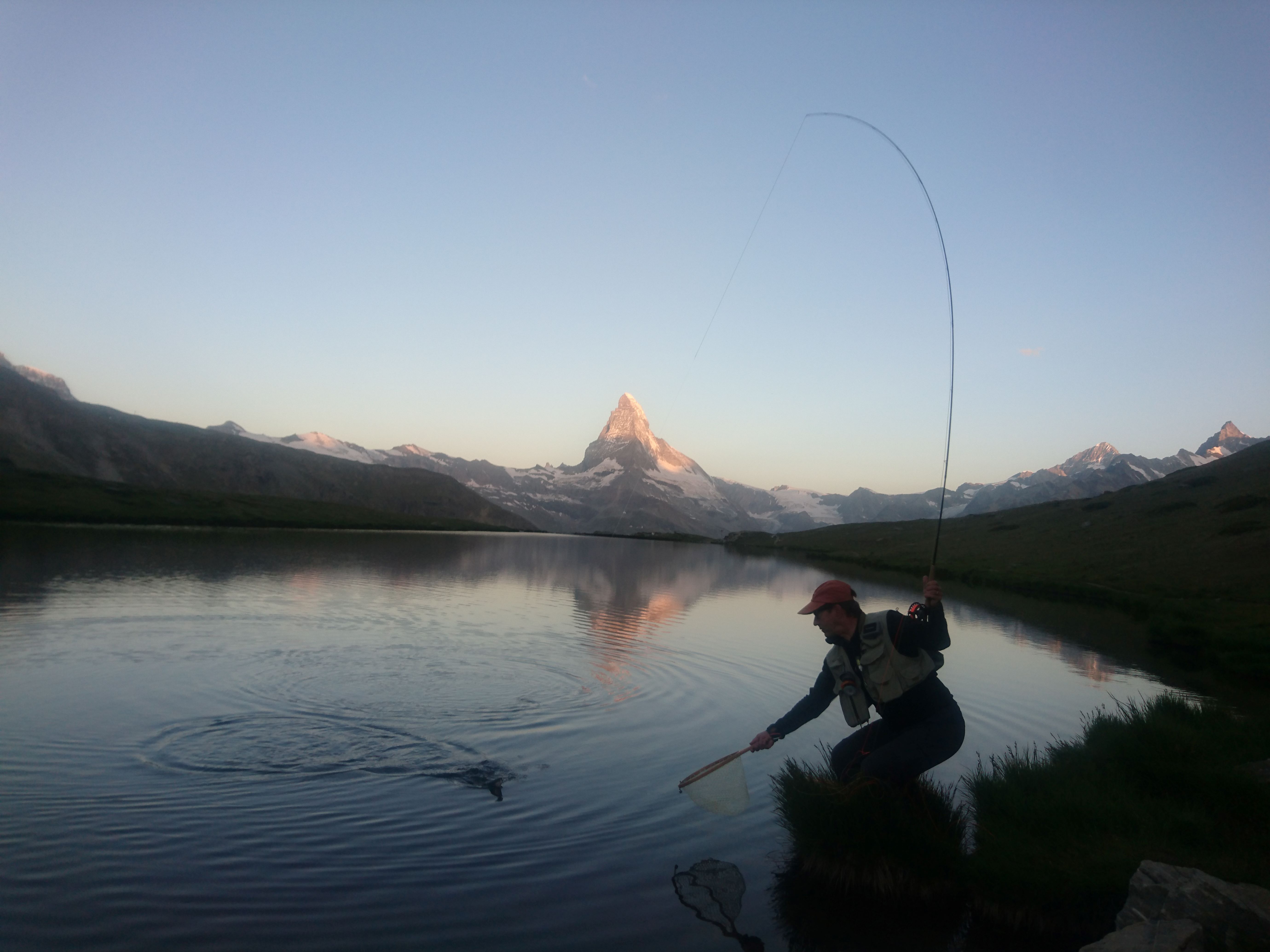 Fly Fishing in Switzerland, landing a trout with a bent rod and the sun just rising on the tip of the Matterhorn
